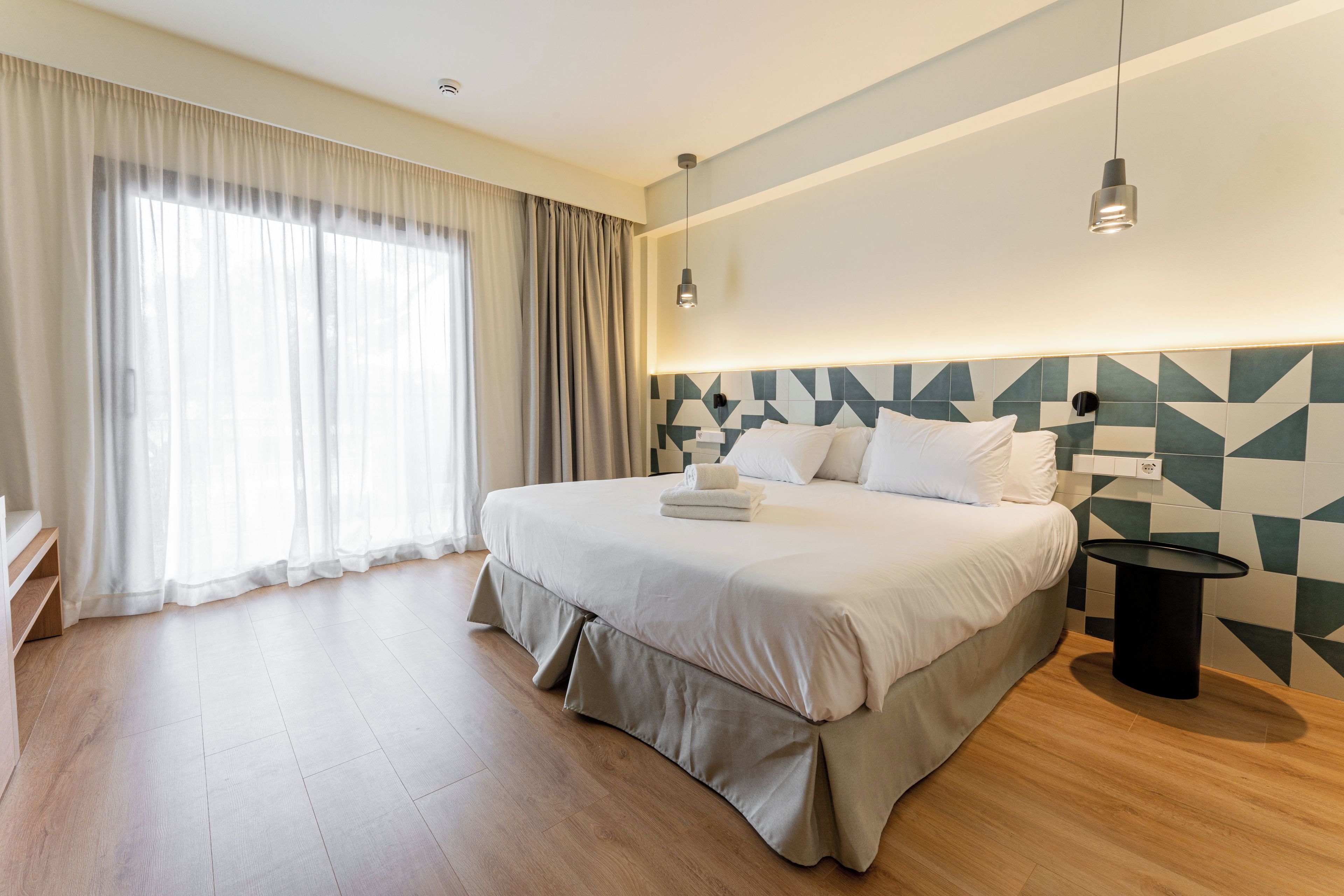 A modern hotel room with a large bed, a bedside table, a wall-mounted lamp, and a window with curtains.