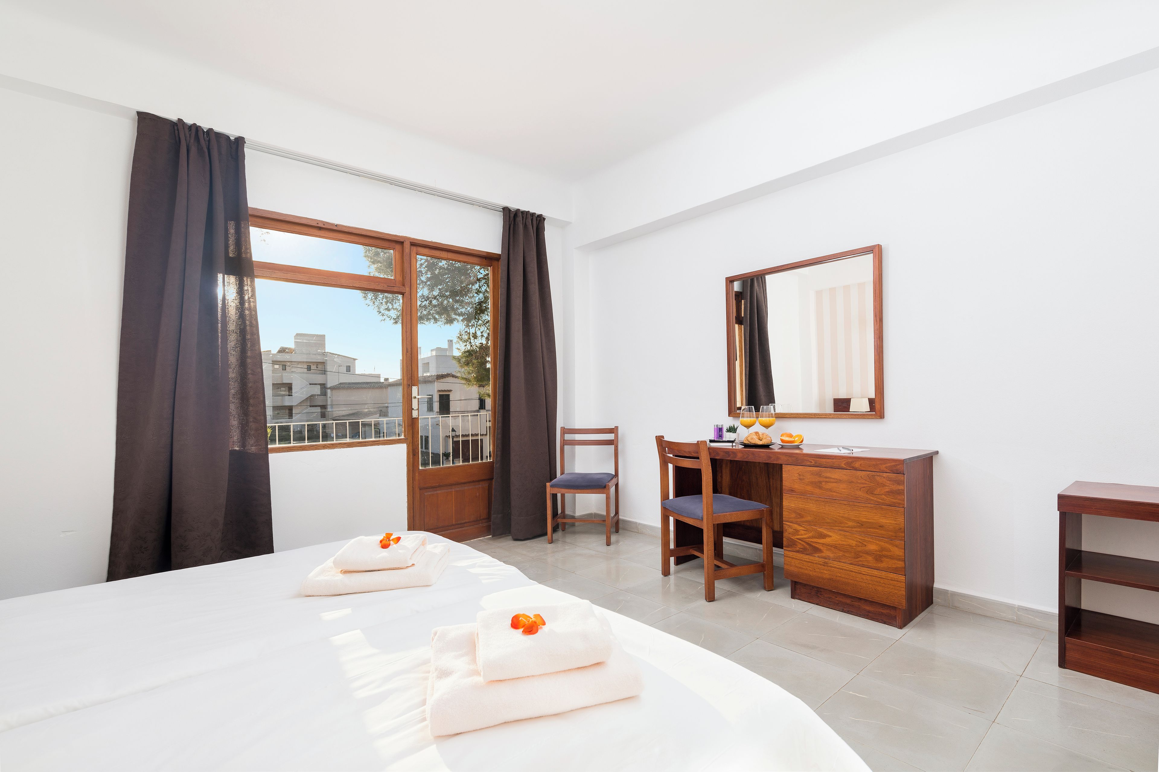 A hotel room with a bed, two chairs, a desk, a dresser, a mirror, and a window with curtains.