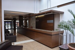 A modern reception area with a wooden reception desk, a seating area with leather sofas, and a large potted plant. - Hotel Presidium Palace (Lovech)