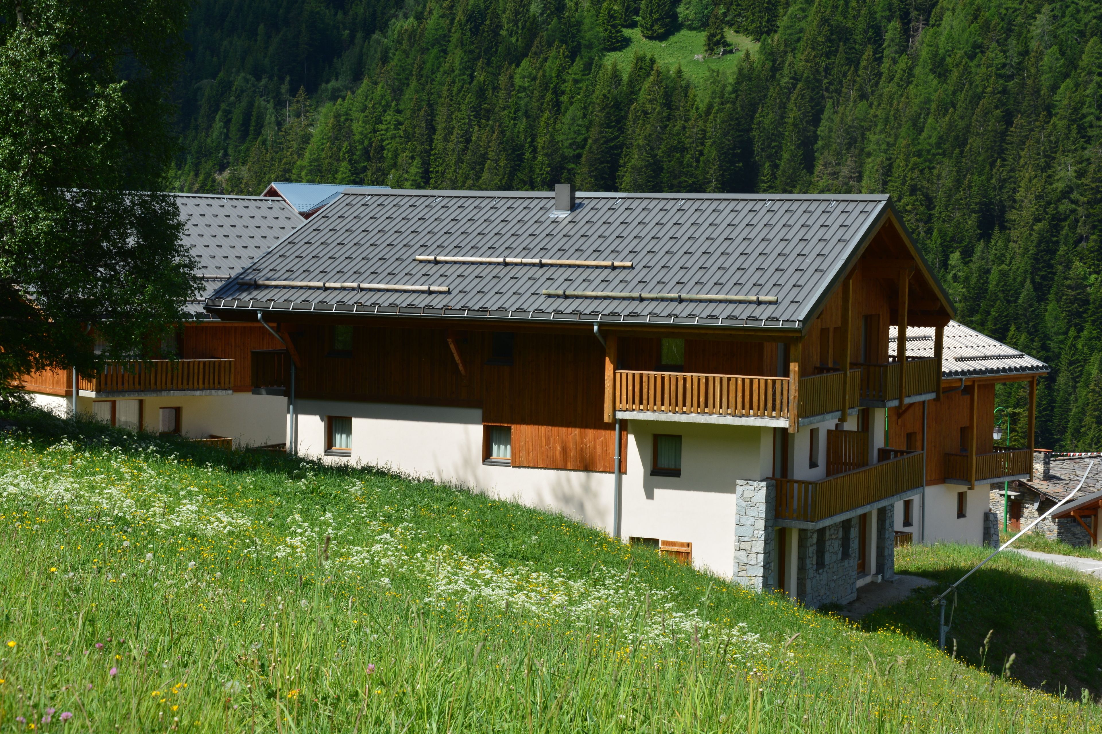 A multi-story wooden chalet with a sloped roof, set on a hillside with a forest backdrop.