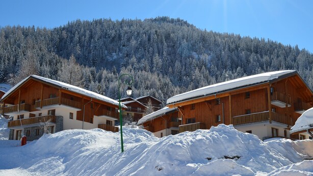 A wooden chalet-style building with balconies, surrounded by snow-covered trees and a mountain in the background. - Vacancéole - Les Chalets de la Ramoure (Modane)