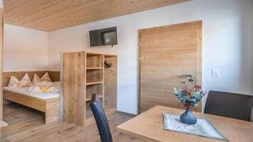 A room with a wooden ceiling, a bed with pillows, a TV mounted on the wall, a wooden door, and a table with a vase of flowers.