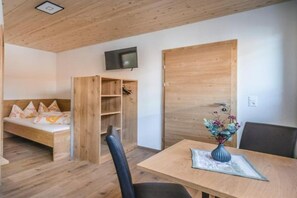 A room with a wooden ceiling, a bed with pillows, a TV mounted on the wall, a wooden door, and a table with a vase of flowers.