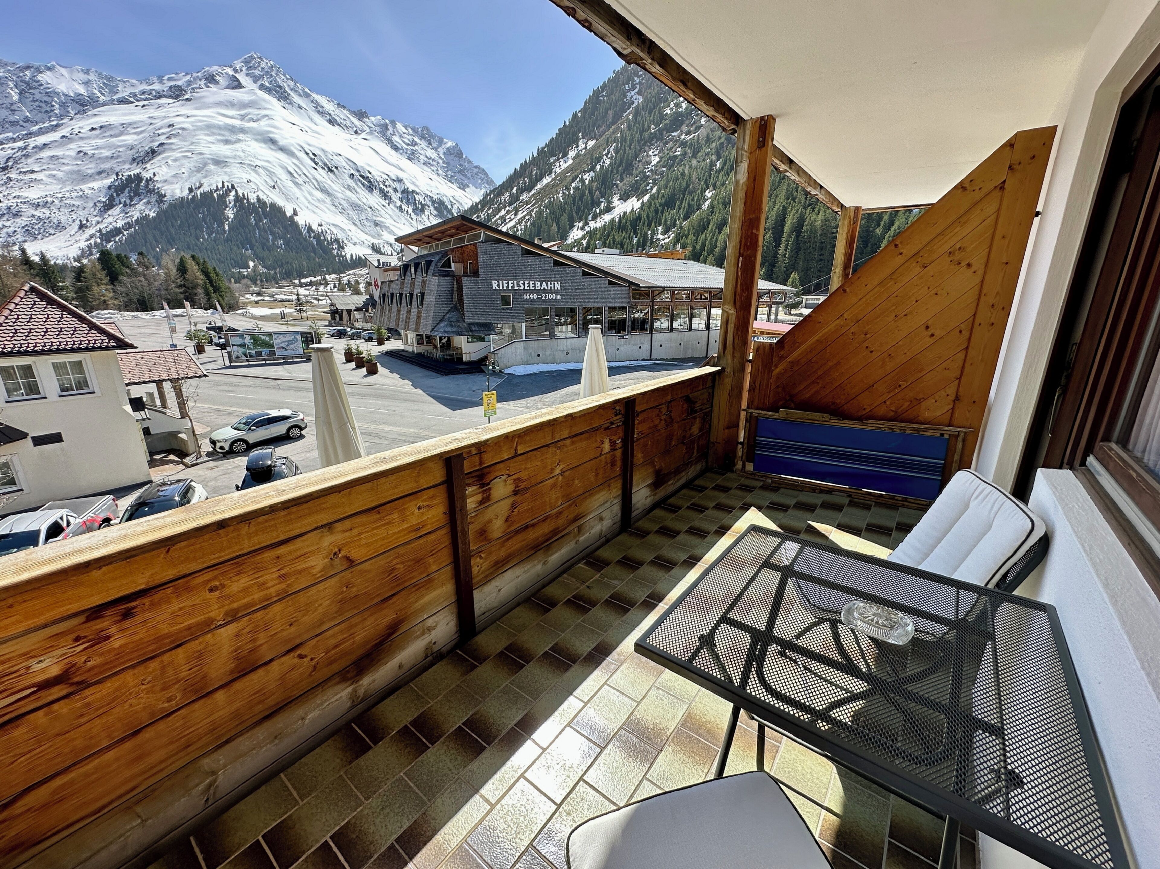 A balcony with a view of snow-capped mountains, a table with chairs, and a building labeled 'Riffelseebahn' in the background.