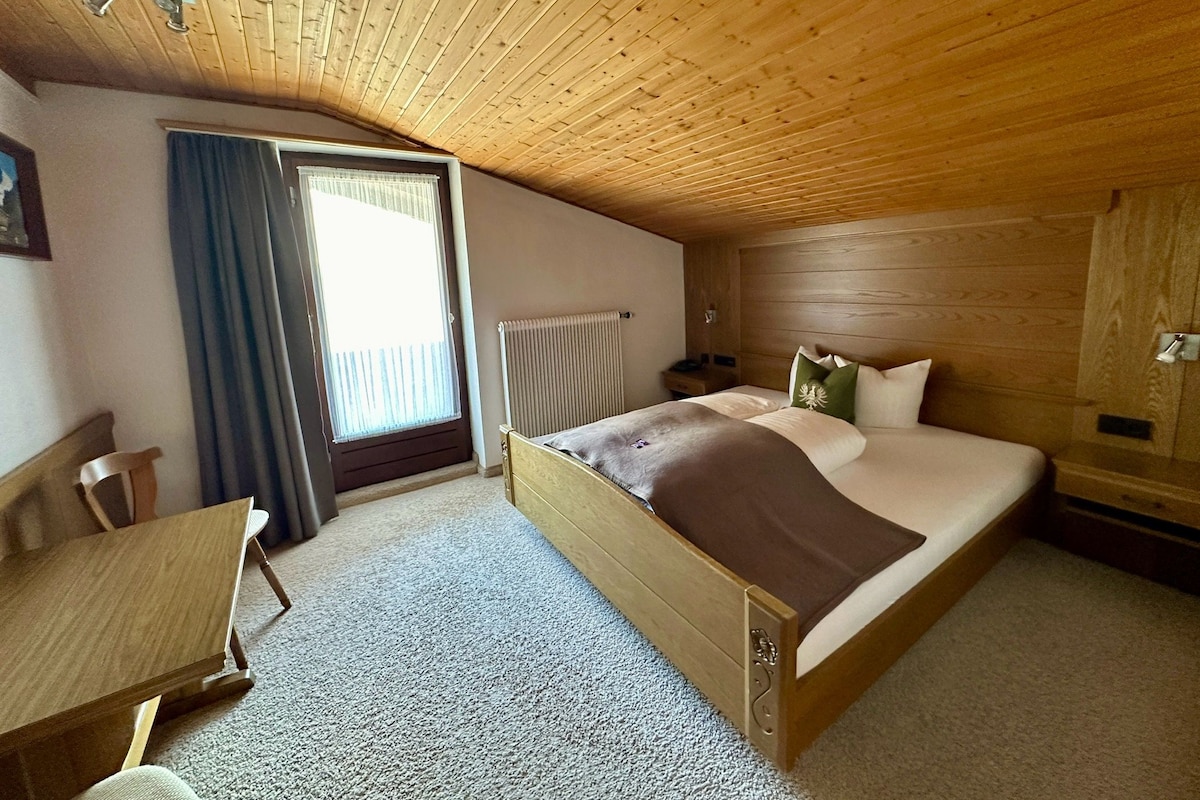 A room with a wooden ceiling, a bed with pillows, a chair, a table, and a window with curtains.