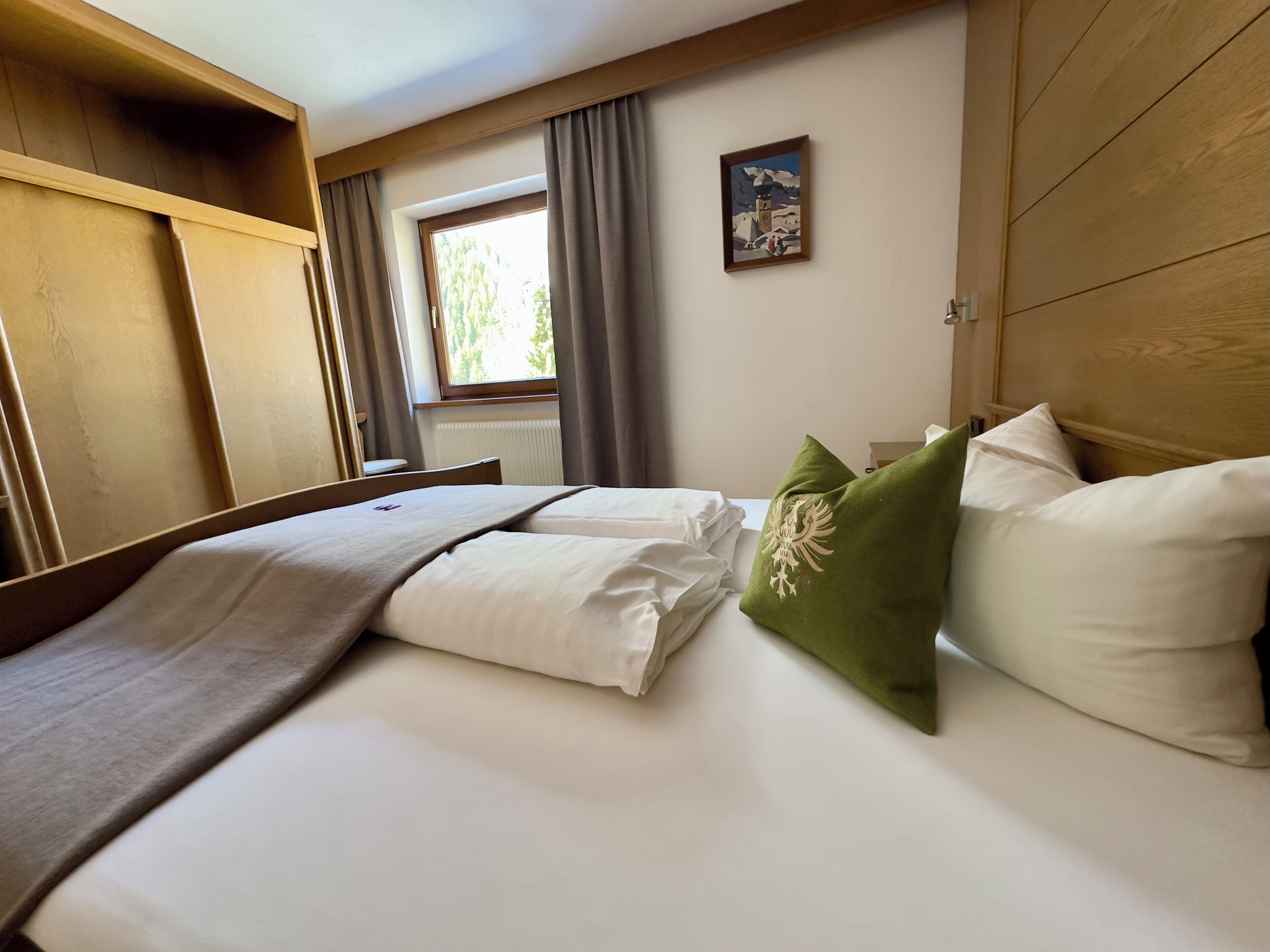 A hotel room with a bed, a wooden headboard, a window with curtains, and a painting on the wall.