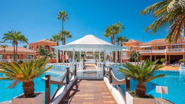 Un espace piscine aménagé dans un complexe hôtelier, avec un kiosque, des palmiers et un ciel d’un bleu limpide.