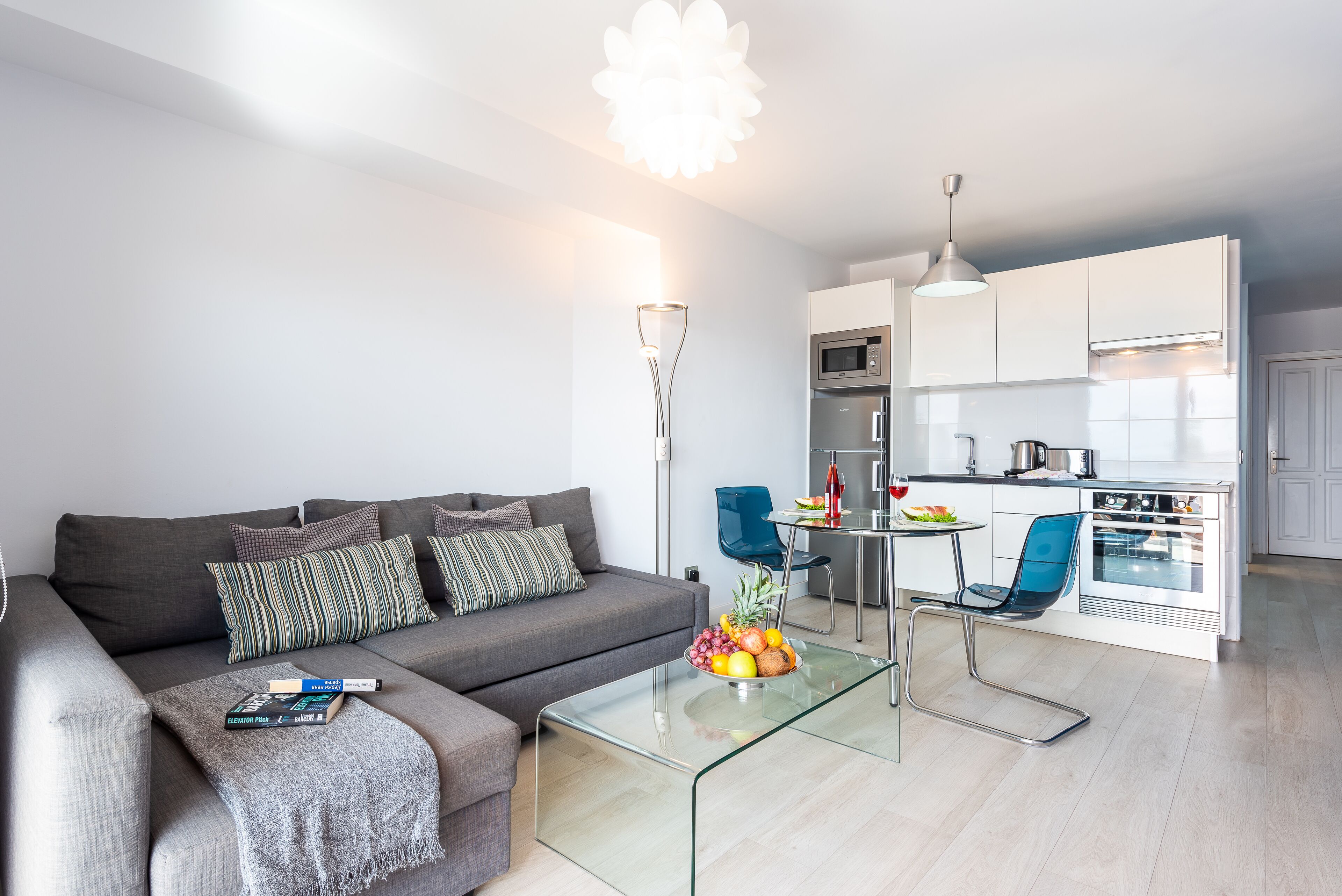 A modern living room with a grey sofa, a glass coffee table with fruit, and a dining area with a white table and blue chairs.