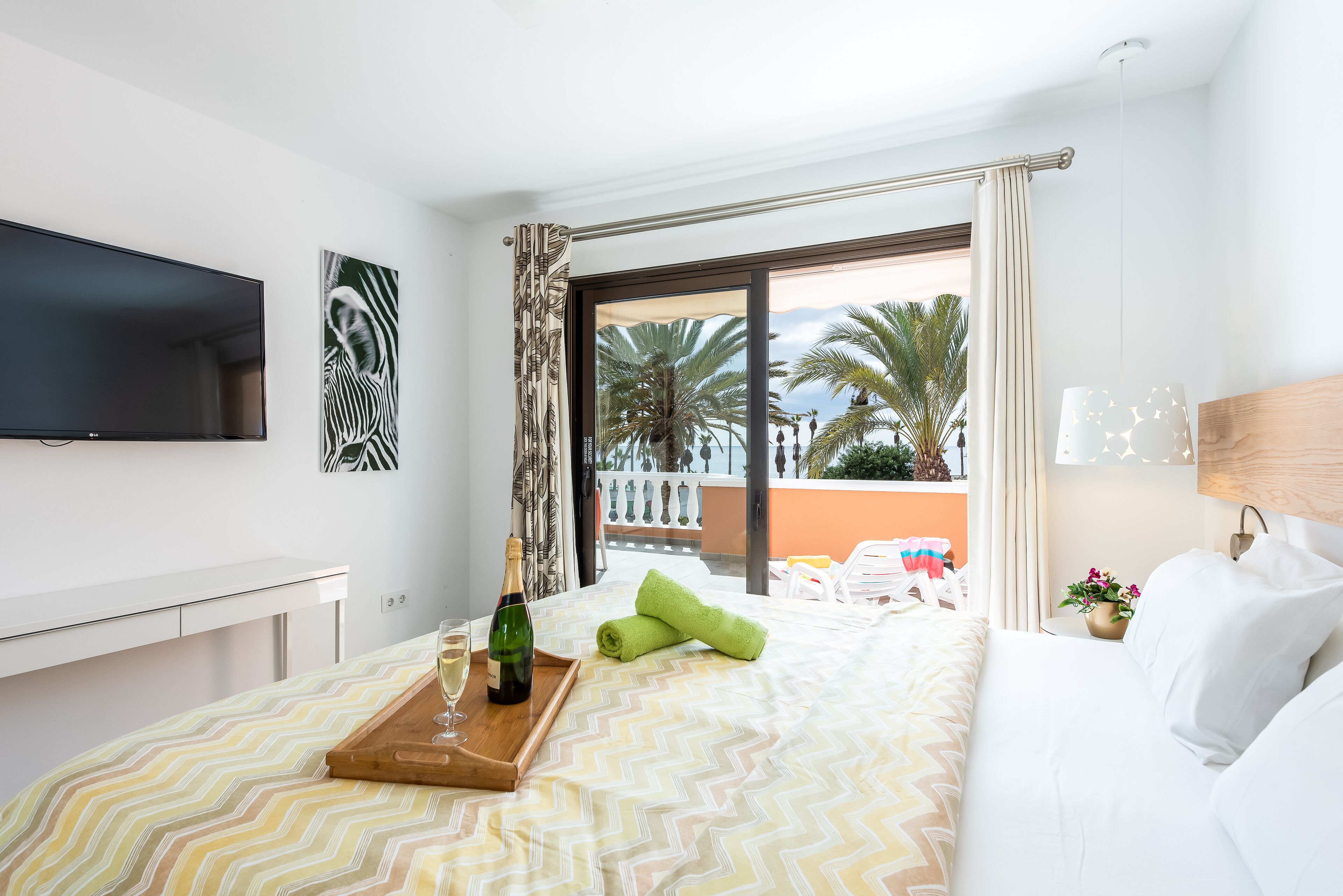 A hotel room with a bed, a TV mounted on the wall, a balcony with a view of palm trees, and a bottle of wine on a tray.