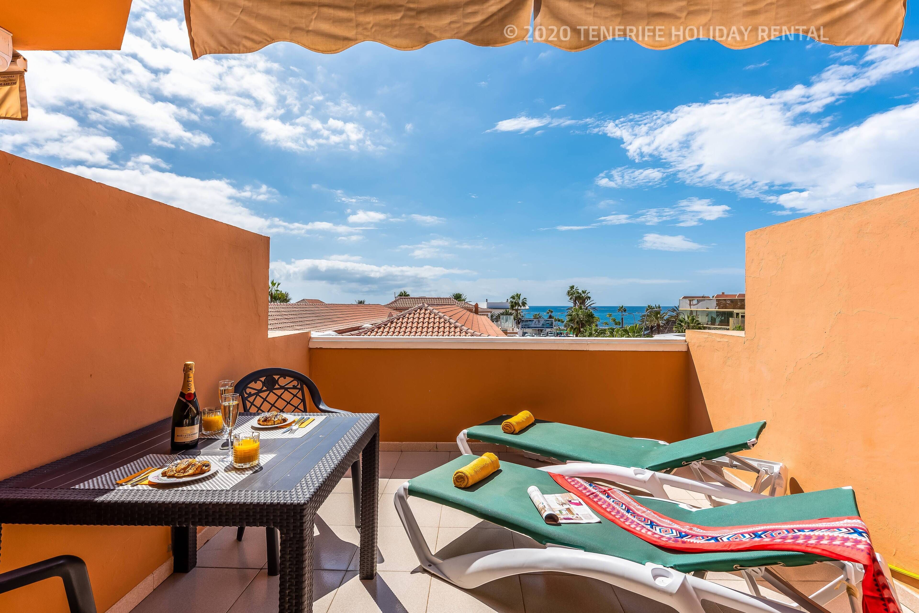 Un balcon avec une table dressée pour deux personnes, une bouteille de champagne et un verre de jus d’orange. On y trouve deux chaises longues avec des serviettes. Le balcon donne sur une zone côtière, avec des bâtiments et des palmiers.