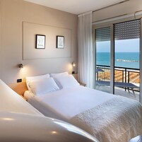 Habitación de hotel con cama, dos cuadros en la pared, una mesita de noche y vista a la playa a través de una puerta corrediza.