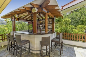 Un área de bar al aire libre con sillas de mimbre, un refrigerador de Coca-Cola y una pérgola de madera con letreros que indican 'Holland' y 'Paris'.