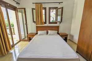 A hotel room with a large bed, two bedside tables, a wooden headboard, and a sliding glass door leading to an outdoor area. - Golden Sun Kokkari (Samos)