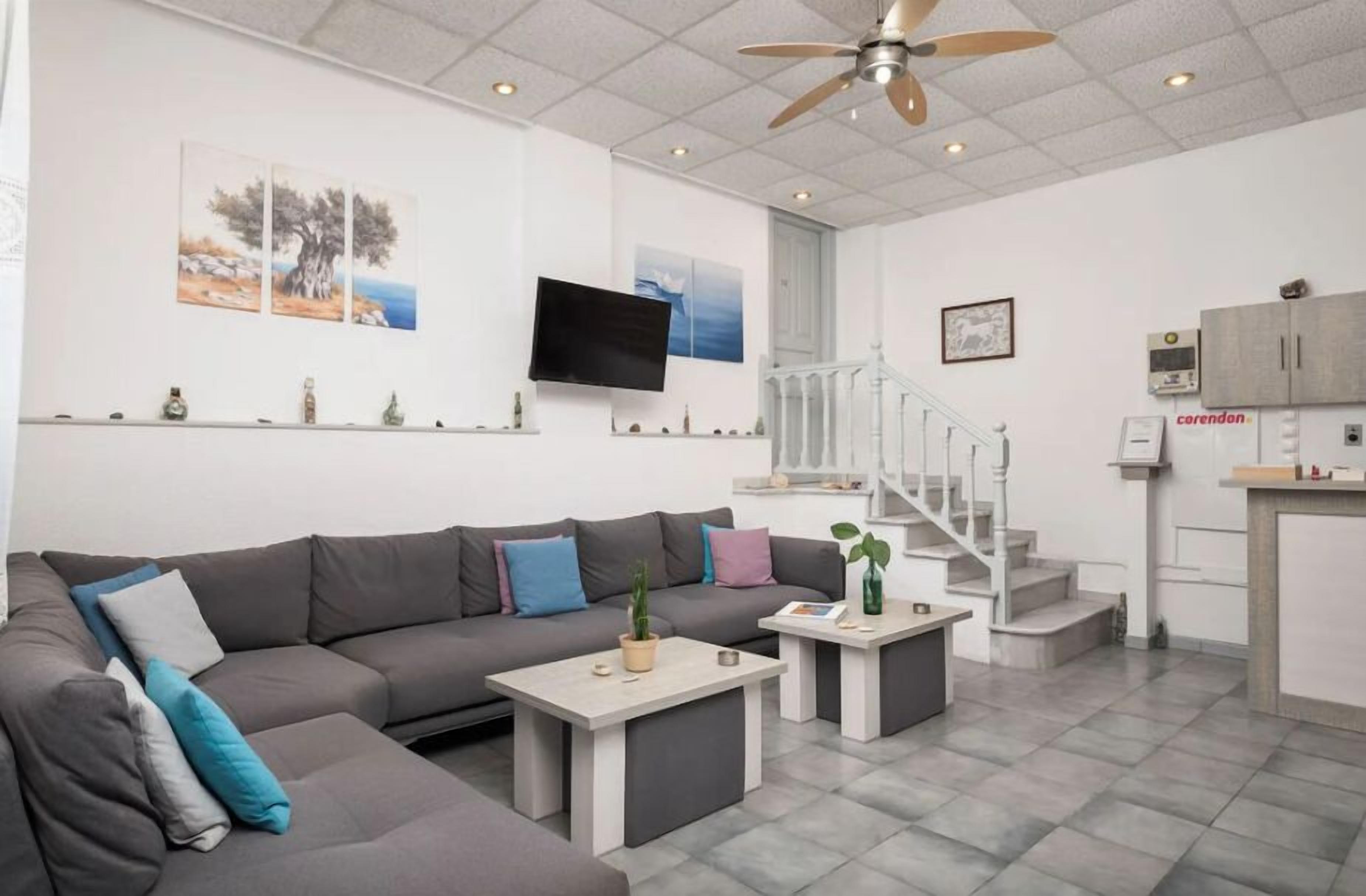 A modern living room with a grey sectional sofa, a flat-screen TV, a ceiling fan, and a staircase leading to a kitchen area.