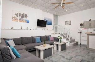 A modern living room with a grey sectional sofa, a flat-screen TV, a ceiling fan, and a staircase leading to a kitchen area.
