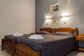 A double bed with a dark-colored bedspread and two folded towels on the bed. There is a wooden nightstand with a lamp and a small table beside the bed. - Golden Sun Kokkari (Samos)