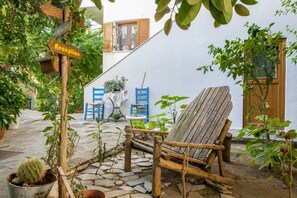 A rustic outdoor seating area with a wooden chair and a table, surrounded by potted plants and a stone pathway. - Golden Sun Kokkari (Samos)