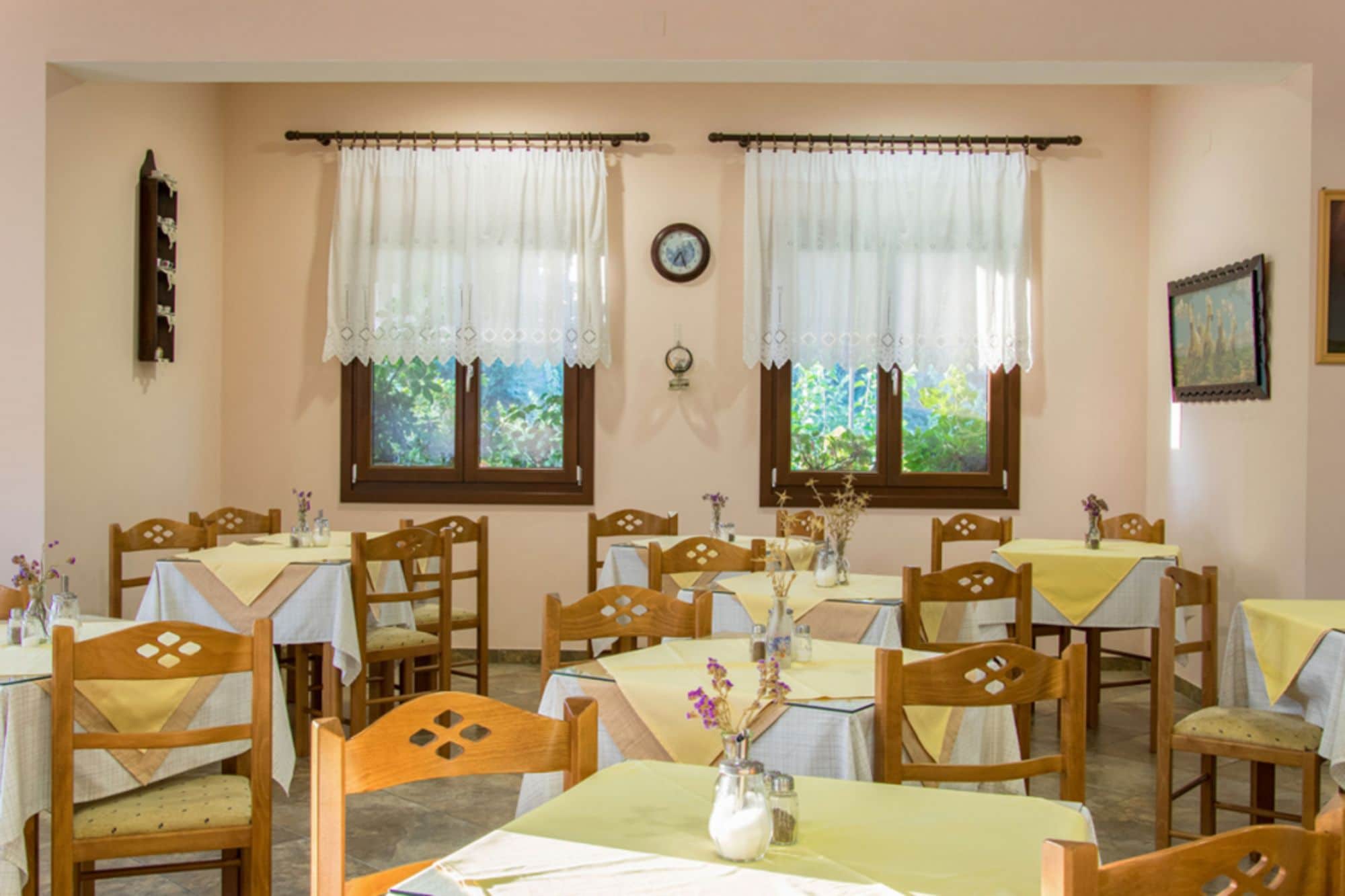 A dining area with multiple tables and chairs, two windows with curtains, and a clock on the wall.