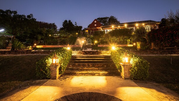 A nighttime view of a building with a red roof, surrounded by a garden with steps leading up to the entrance, and illuminated by street lamps. - Phu Vara (Chiang Dao)
