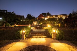 Een nachtzicht van een gebouw met een rood dak, omgeven door een tuin met trappen naar de ingang, verlicht door straatlantaarns.