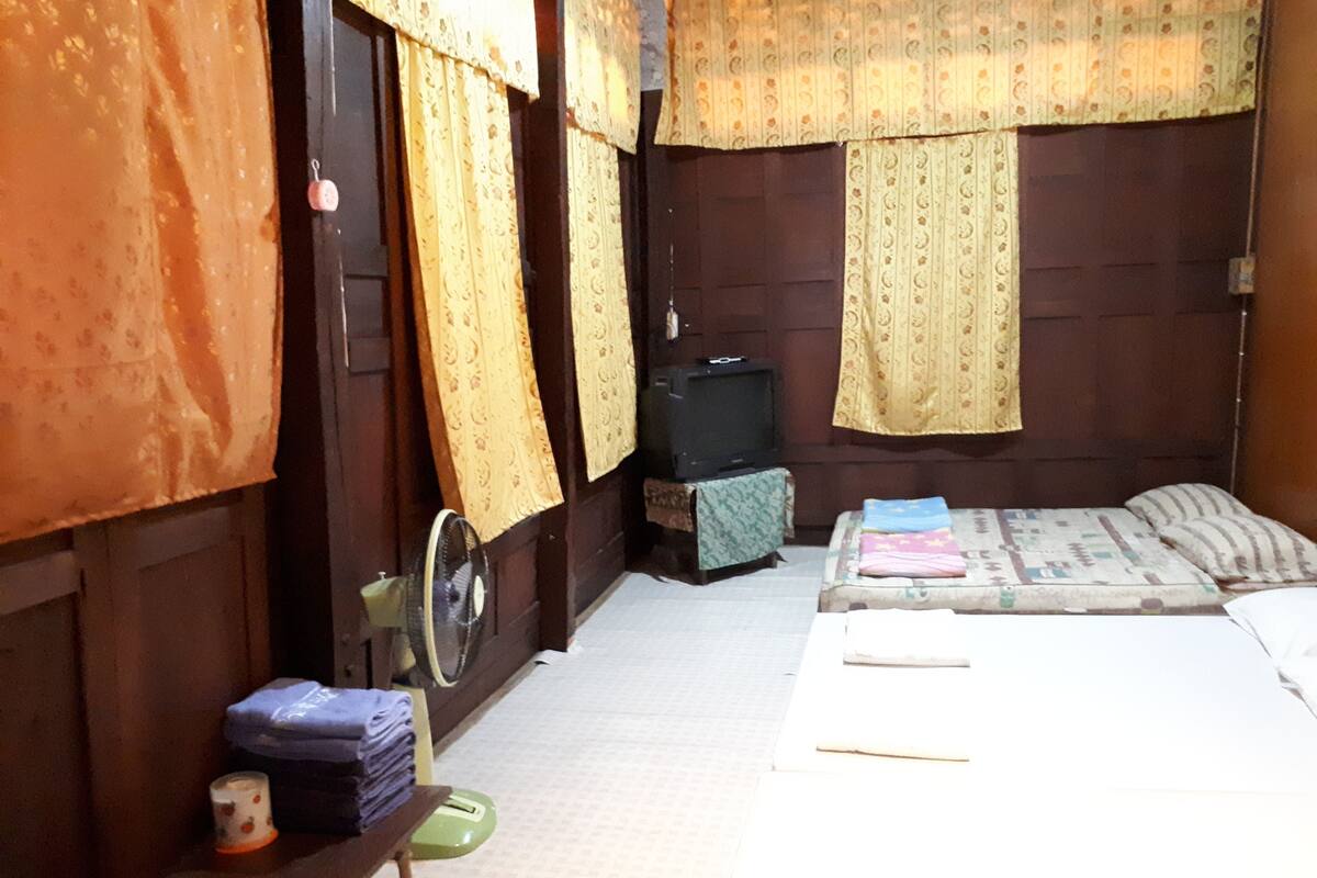 A small, dimly lit room with a single bed, a small table with a fan, and a television on a stand.