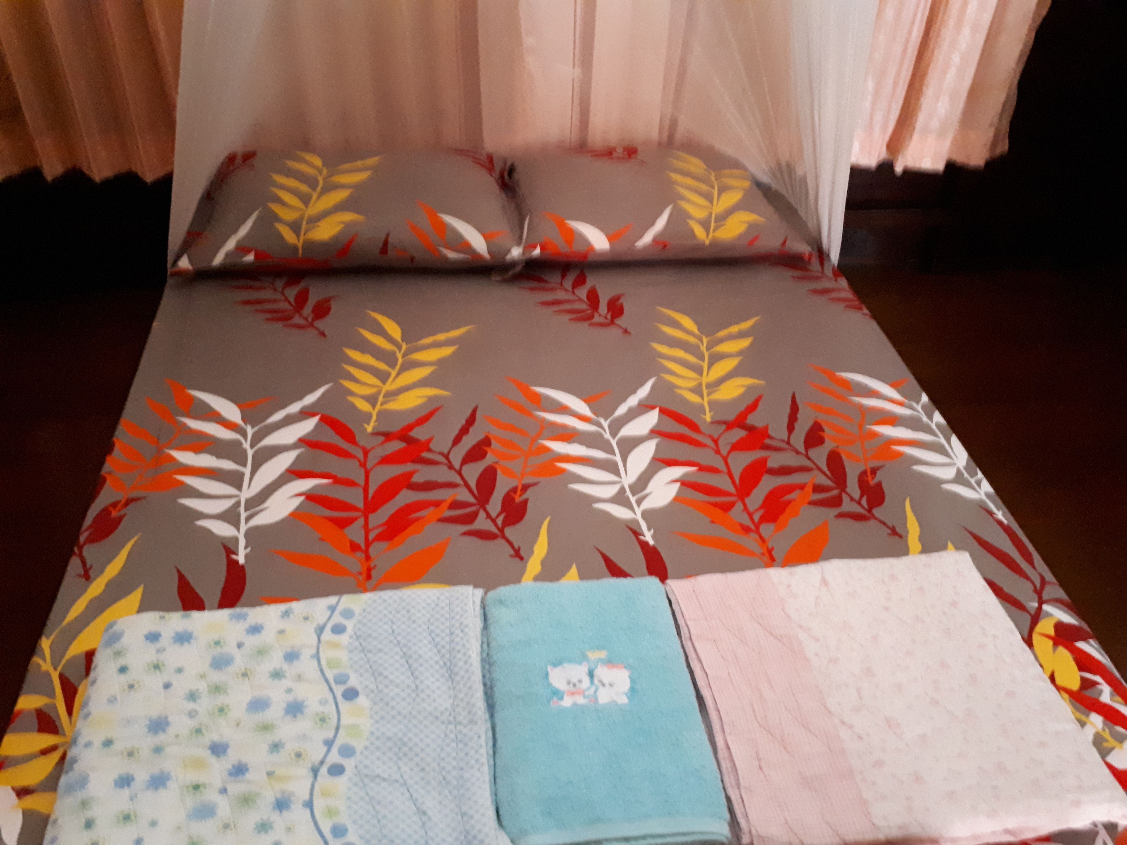 Una cama con un edredón y almohadas estampadas con motivos de hojas, una mesita de noche con toallas dobladas y una mosquitera sobre la cama.