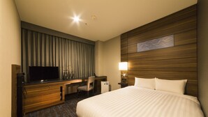 A hotel room with a large bed, a desk with a television, a chair, a nightstand with a lamp, and a wall-mounted artwork.