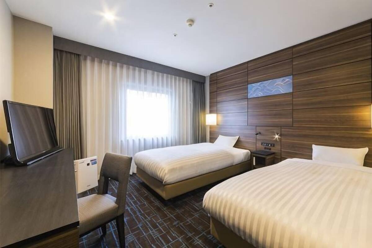 Habitació d'hotel amb dos llits, escriptori, cadira, televisió i finestra amb cortines.