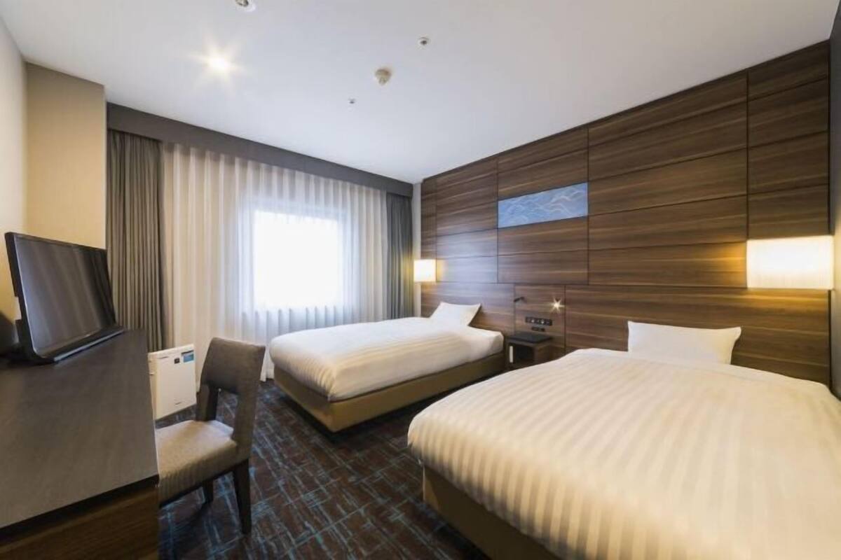 Habitació d'hotel amb dos llits, una escriptori, una cadira, un televisor i una finestra gran amb cortines.