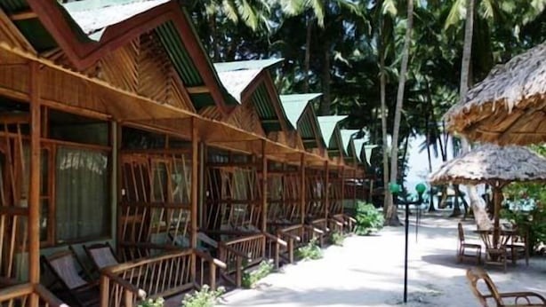 A row of wooden bungalows with thatched roofs, arranged in a line along a sandy path, surrounded by palm trees. - Aquays Hotels & Resorts (Port Blair)