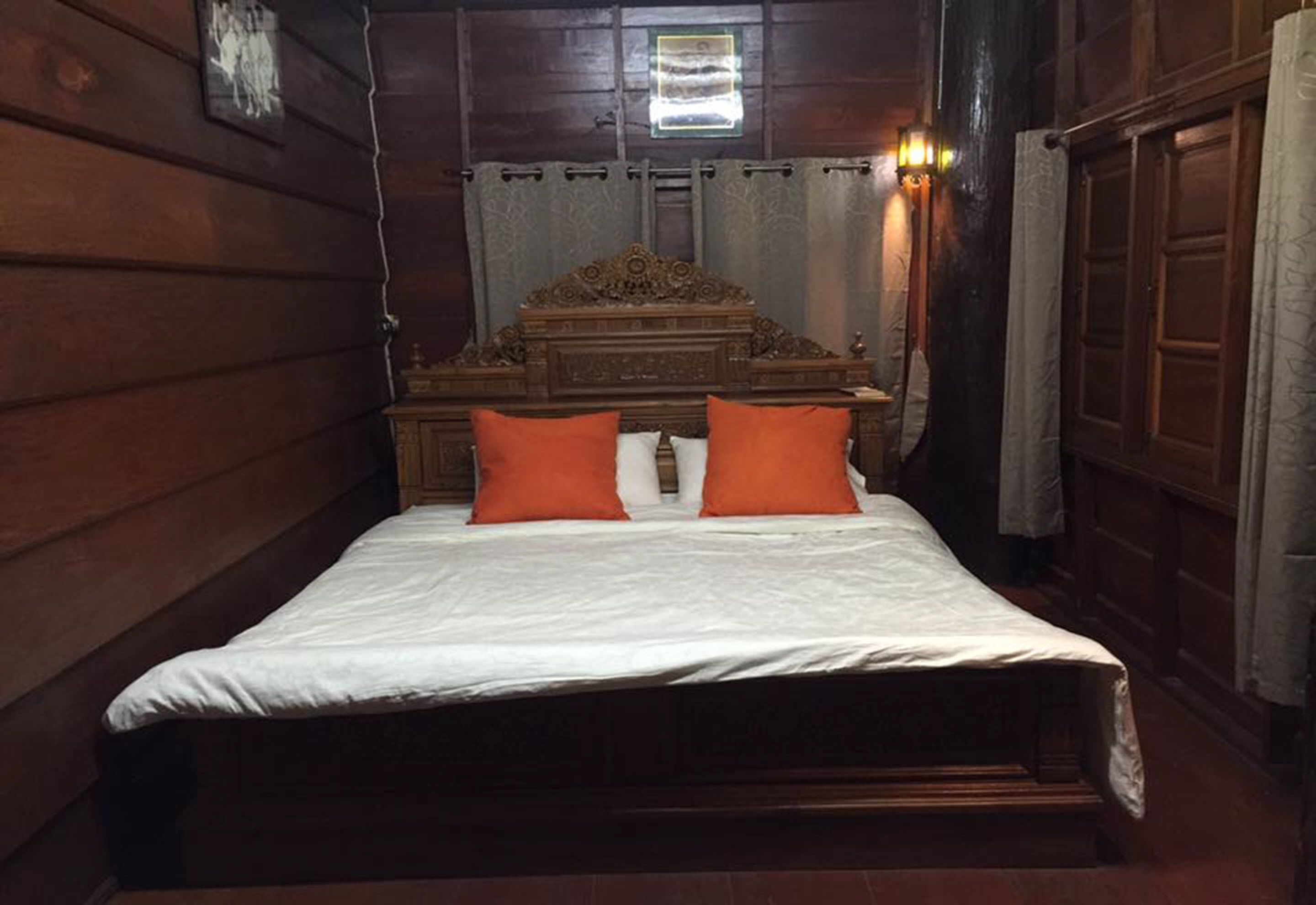 A wooden room with a bed, two orange pillows, a wooden headboard, and a window with curtains.