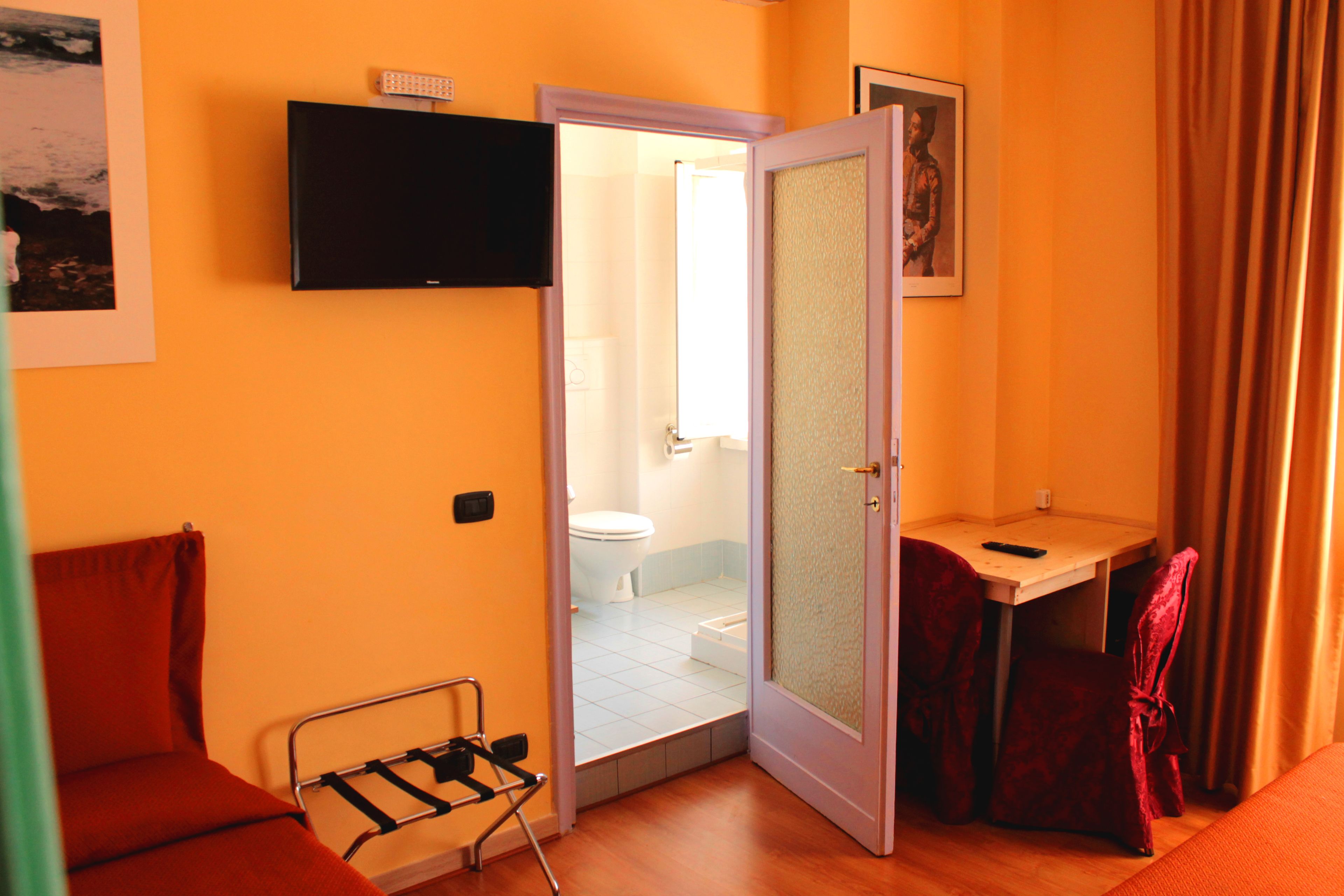 A compact hotel room with a bed, a small desk, a chair, a television, and a bathroom visible through an open door.