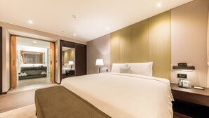 A modern hotel room with a large bed, a bedside table with a phone and a clock, a desk with a lamp, and a bathroom visible through an open door. - Howard Johnson by Wyndham Incheon Airport (Incheon)