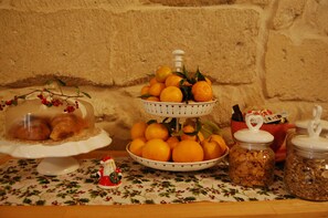 A table with a floral tablecloth, a three-tiered fruit bowl with oranges, a glass jar of cereal, and a glass jar of nuts. - Kabala b&b (Lecce)