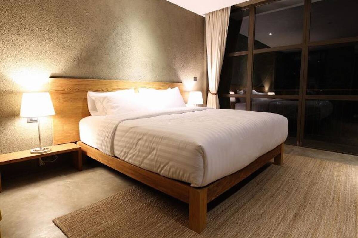 A hotel room with a wooden bed, two bedside lamps, a large window with curtains, and a textured wall.