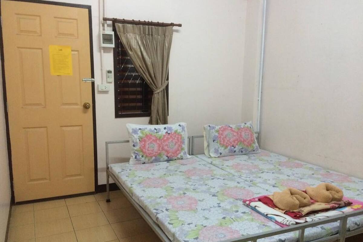 Una habitación pequeña con cama individual, colcha estampada, dos almohadas, puerta de madera, ventana con cortinas y suelo de baldosas.