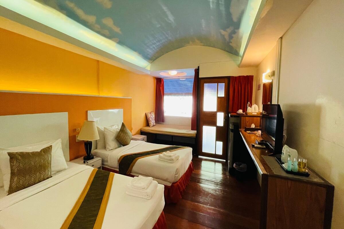 A hotel room with two beds, a desk with a television, and a view of the sky through a window.
