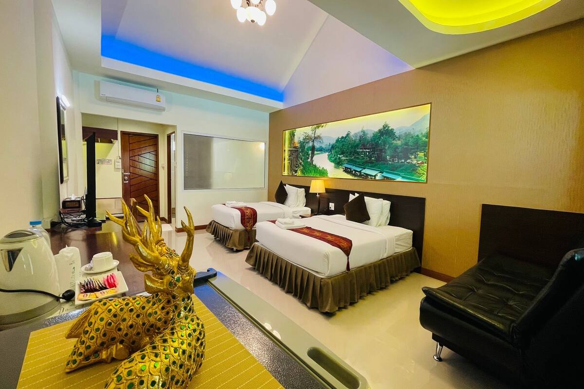 A hotel room with two beds, a sofa, a decorative golden deer statue, a painting of a landscape, and a small table with a teapot and cups.