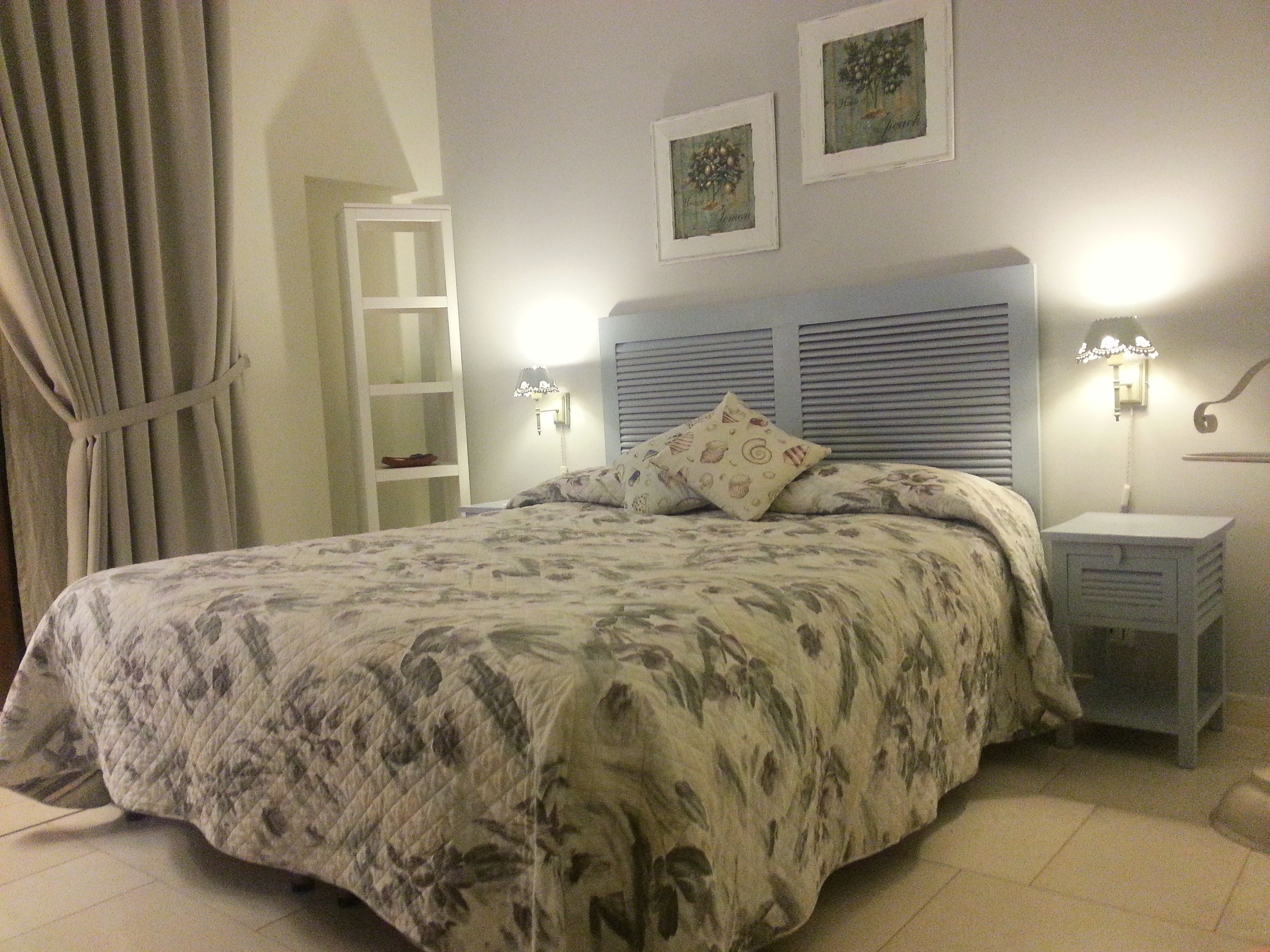 Quarto com cama, mesa de cabeceira, abajur, prateleira e cortinas.