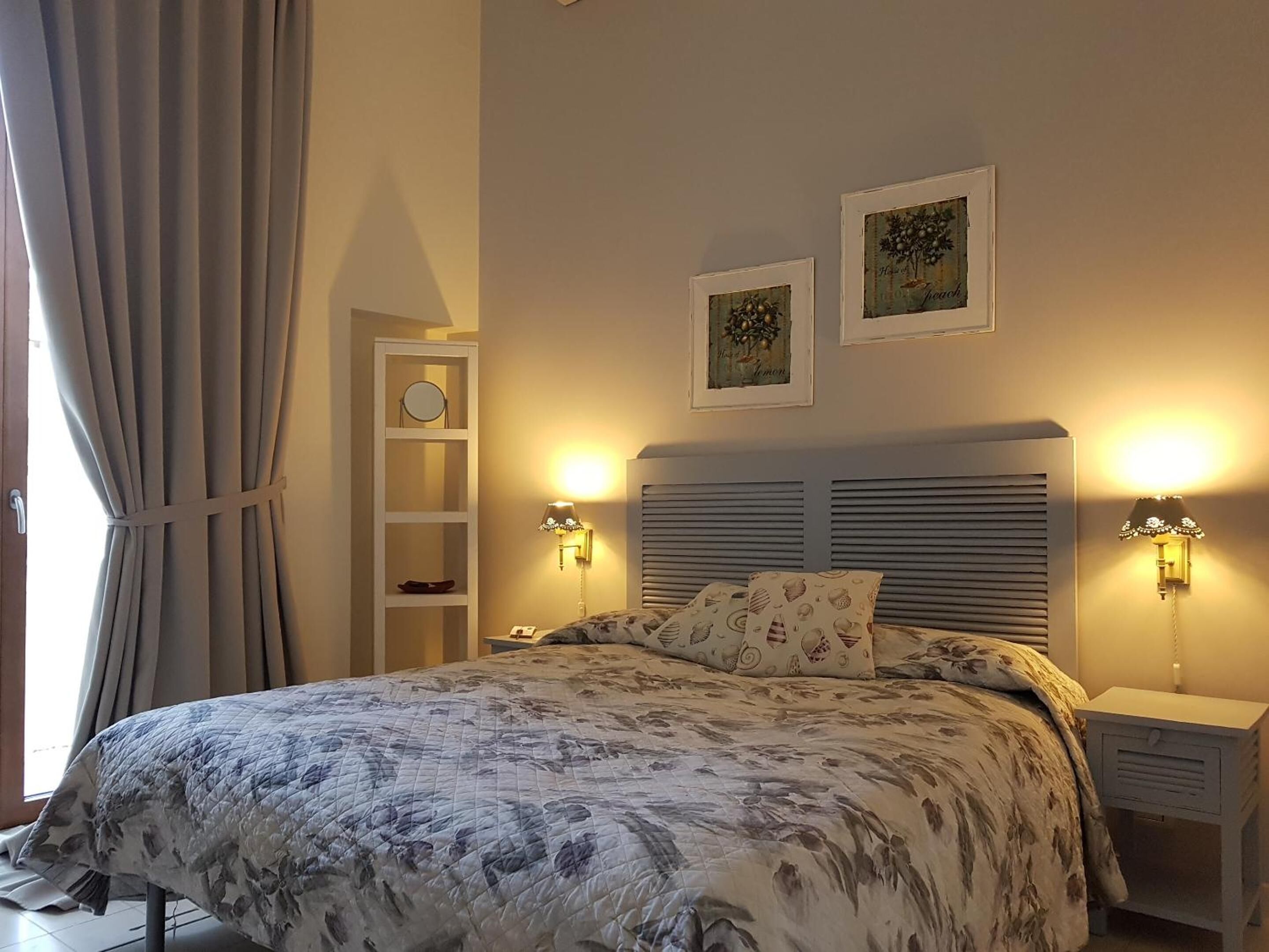 A bedroom with a bed, two wall-mounted lamps, a nightstand, a shelf with a mirror, and two framed pictures on the wall.