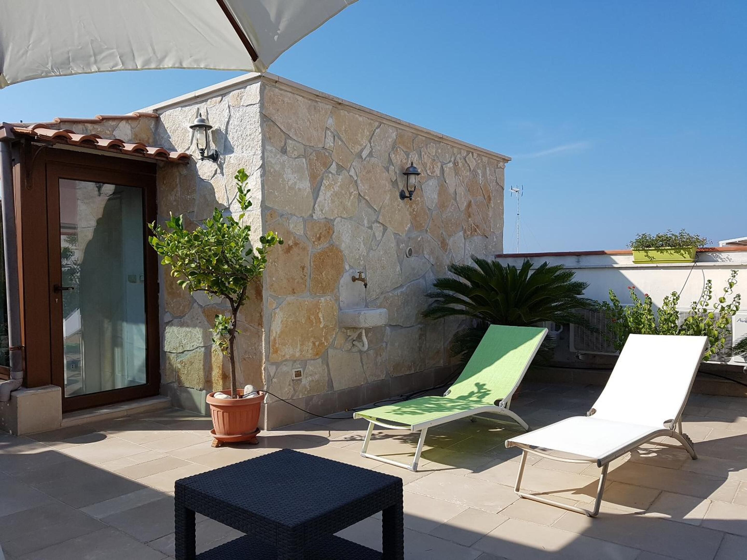 Patio esterno luminoso con pareti in pietra, ingresso con porta in vetro, un albero in vaso, una poltrona da relax verde e una bianca, e un tavolino.