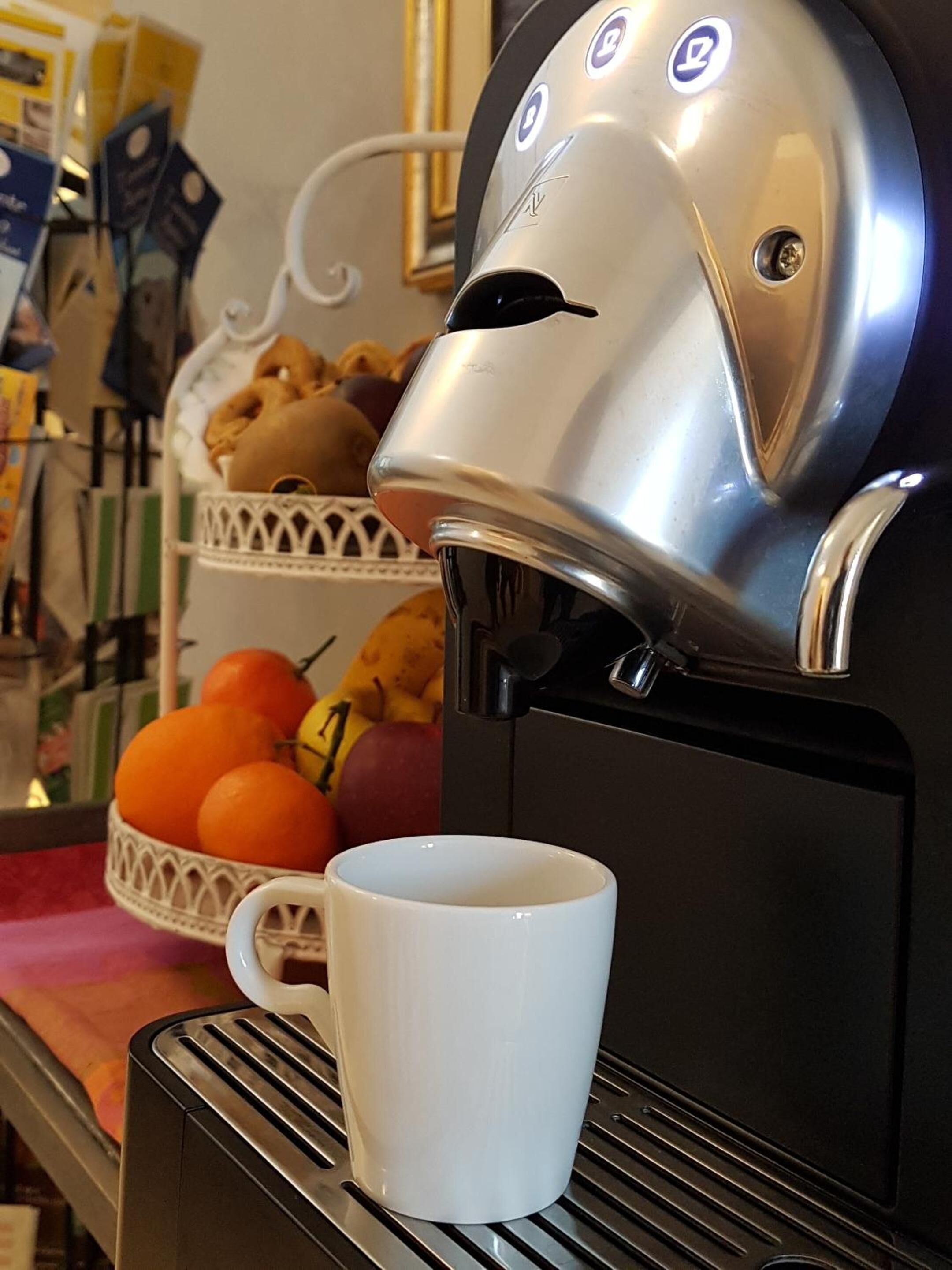A coffee machine with a white mug on its drip tray, a basket of oranges, and a basket of pastries.