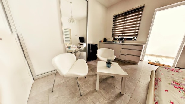 Een moderne slaapkamer met een bed, een klein tafeltje, twee stoelen, een spiegel, een kitchenette en een raam met jaloezieën.