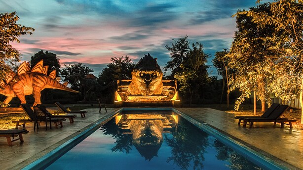 Una piscina con una escultura de dinosaurio y una estatua de león en el centro, rodeada de sillas de descanso y árboles.