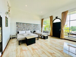 A spacious bedroom with a large bed, a bench, and a small table. There is a window with curtains and a view of greenery outside. - Yoko River Kwai Resort (Sai Yok)