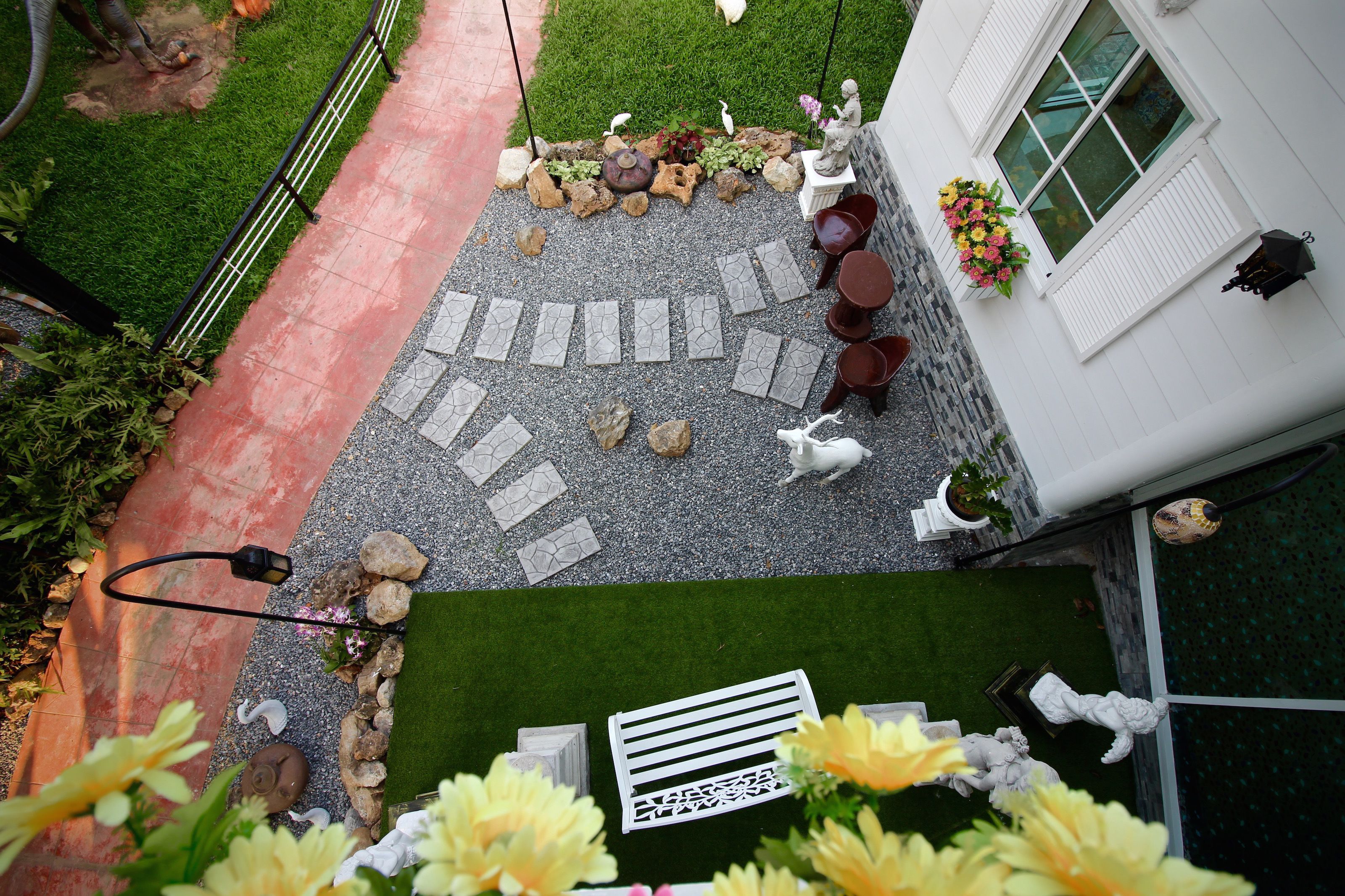 A garden patio with a stone pathway, seating area, and decorative elements like statues and flowers.