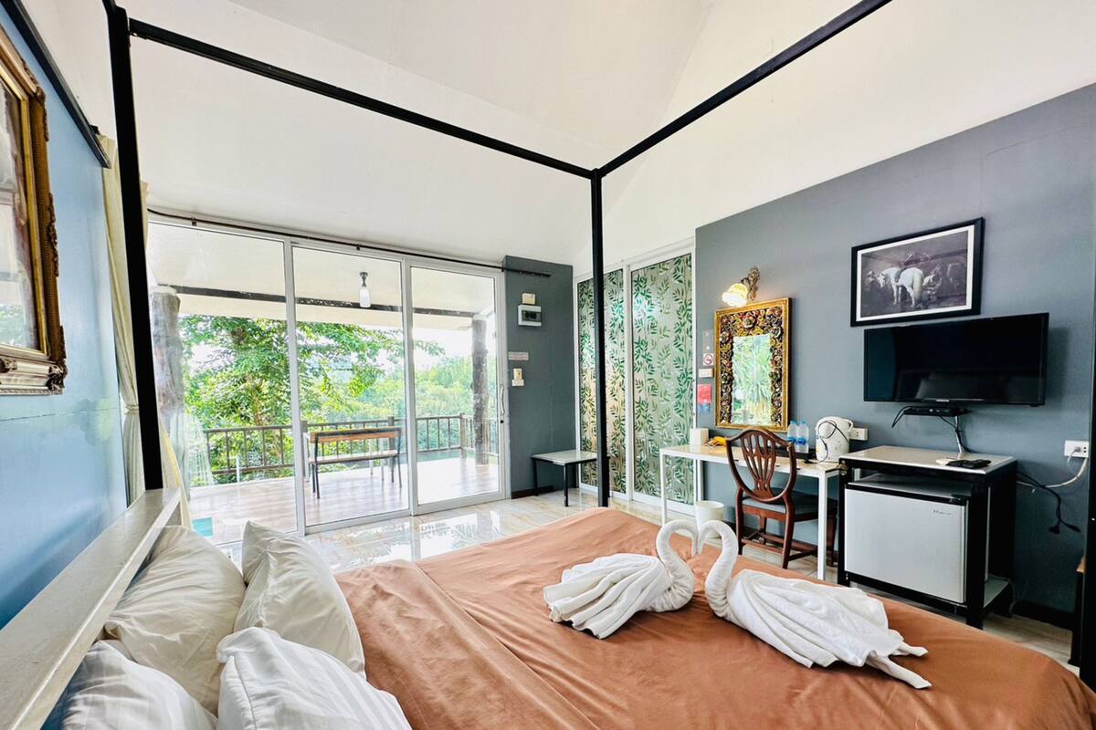 A hotel room with a bed, a desk, a chair, a television, and a balcony with a view of greenery.