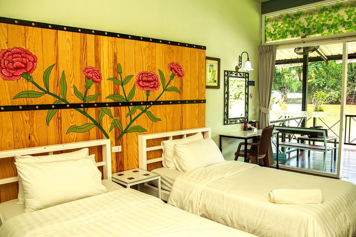 A hotel room with two beds, a wooden headboard with floral artwork, a small table, and a view of greenery outside.