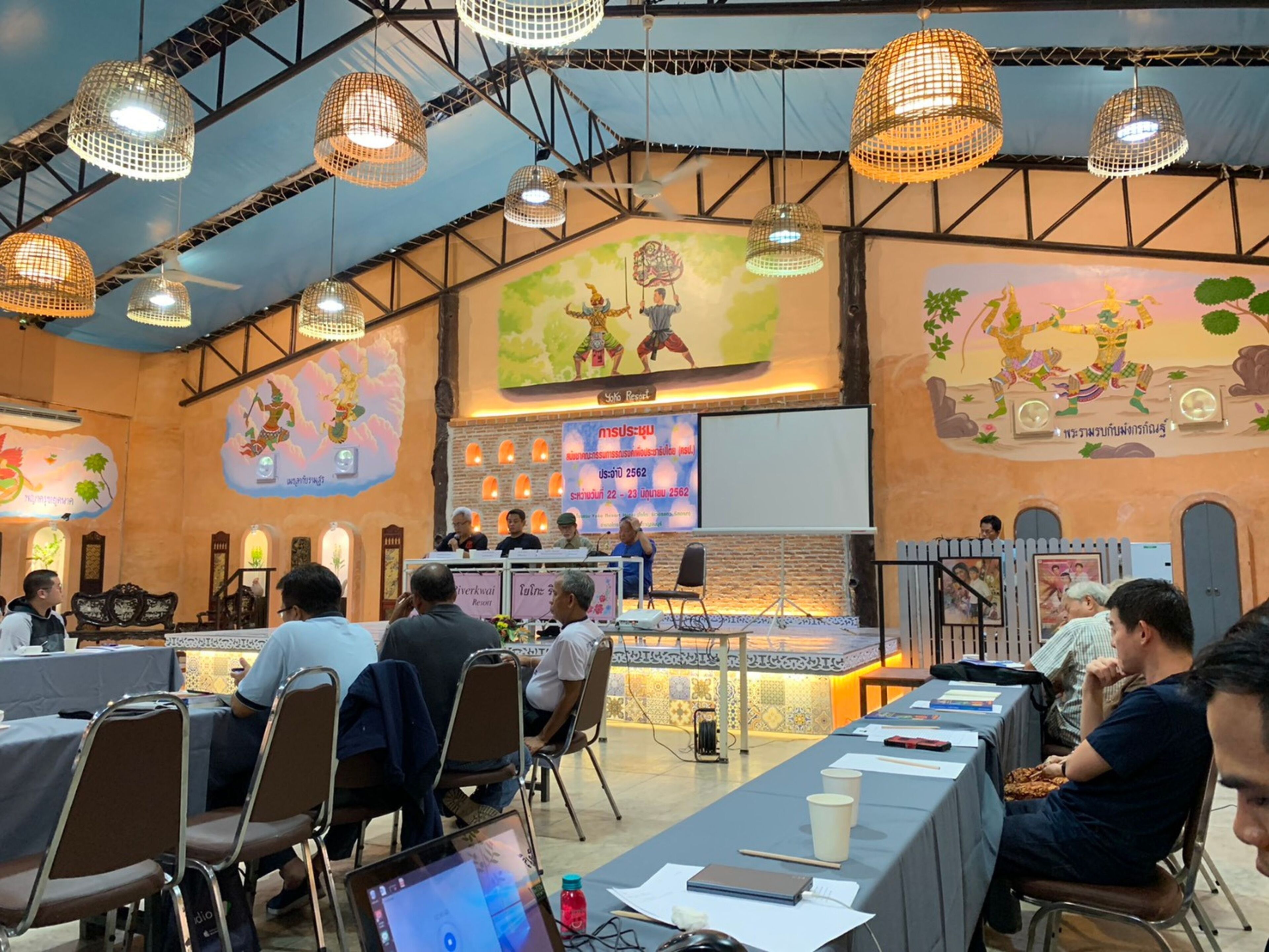 Un salón de conferencias con escenario, asistentes sentados en mesas y murales decorativos en las paredes.