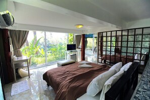 A hotel room with a large bed, a desk, a TV, and a balcony with a view of greenery. - Yoko River Kwai Resort (Sai Yok)